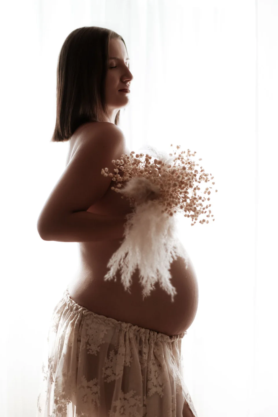 Babybauchporträt mit Trockenblumen im weichen Fensterlicht